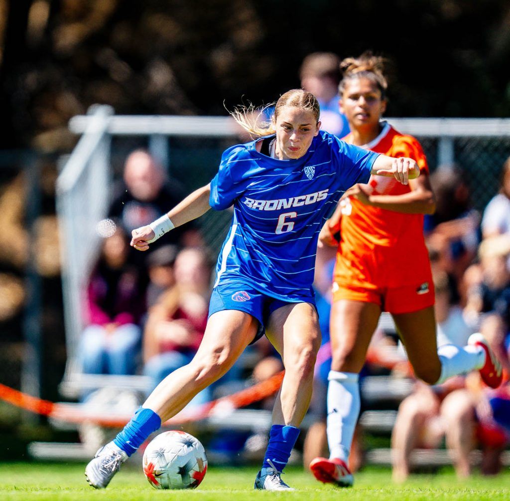 A soccer player in a blue uniform with the number 6 controls the ball while being pursued by an opponent in an orange jersey during a match.