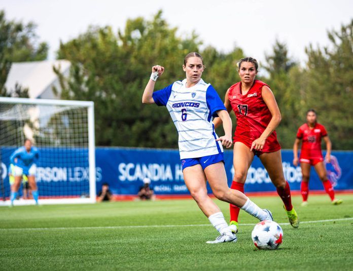Two soccer players compete for the ball on a field, one in a blue and white uniform and the other in red, with a goal and spectators in the background.