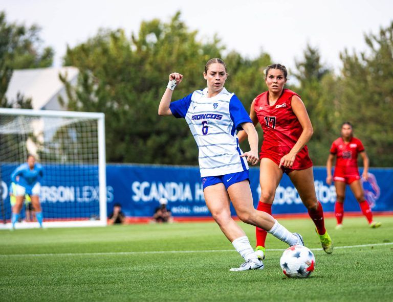 Two soccer players compete for the ball on a field, one in a blue and white uniform and the other in red, with a goal and spectators in the background.