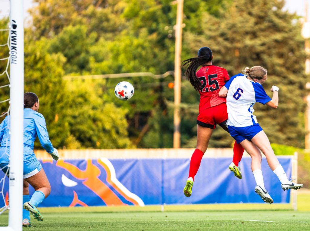 Two players leap in mid-air to challenge for the ball near the goal during a soccer match.